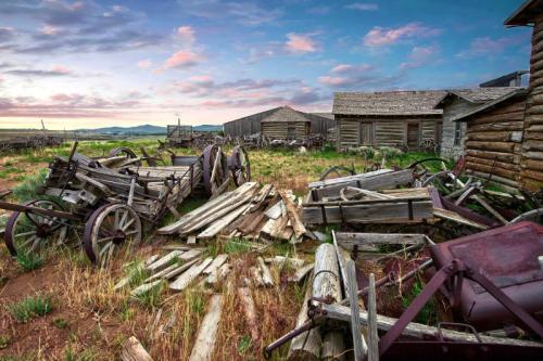 Abandoned farmstead with old wooden structures and broken wagons, set against a colorful sky at dusk.