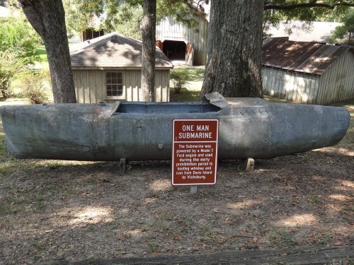 A one-man submarine displayed outdoors, with a sign detailing its history, surrounded by trees and buildings.