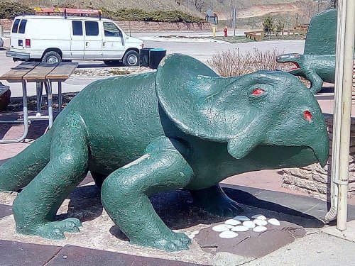 A green, dinosaur-like sculpture with an elephantine head, positioned near a picnic area and a white van in the background.