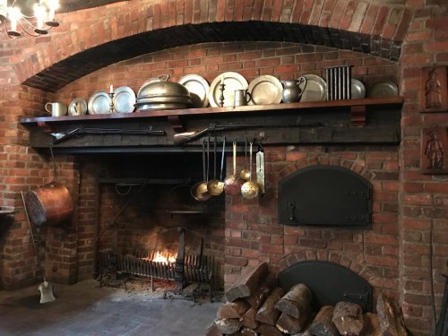 A rustic kitchen with a brick wall, a fireplace, and shelves displaying various metal cookware and utensils.