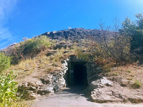 A rocky hillside with a dark cave entrance, surrounded by sparse vegetation and a clear blue sky.