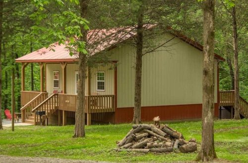 A small cabin with a red roof, surrounded by trees, and a woodpile in front on a grassy area.