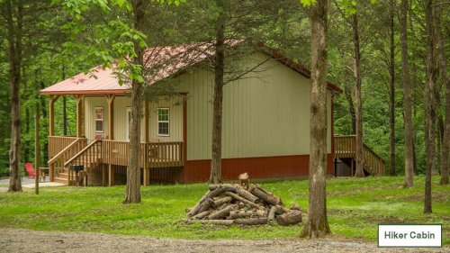 A cozy cabin with a red roof surrounded by trees, featuring a porch and a stack of firewood in front.