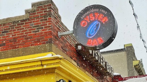 A neon sign reading "Oyster Bar" hangs above a brick building, with snow falling around it.