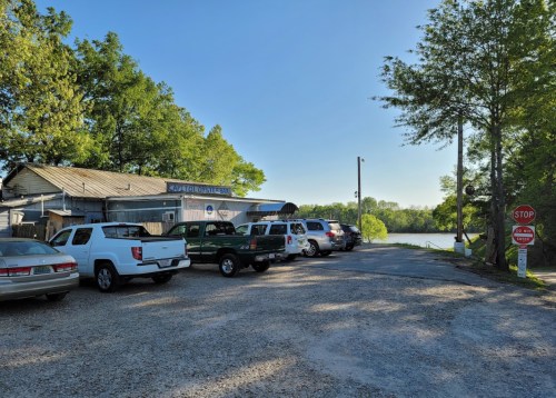 A gravel parking area with several vehicles near a rustic building by a calm river, surrounded by trees.