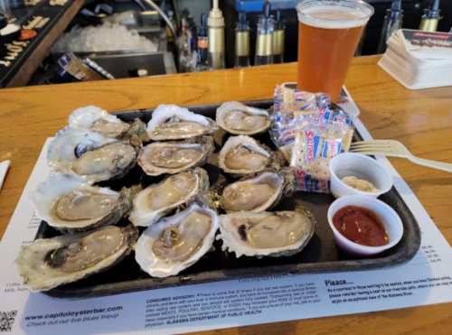 A platter of fresh oysters on ice, accompanied by a beer, cocktail sauce, and condiments on a wooden bar.