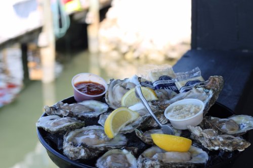 A platter of fresh oysters with lemon wedges and dipping sauces, set against a waterfront background.