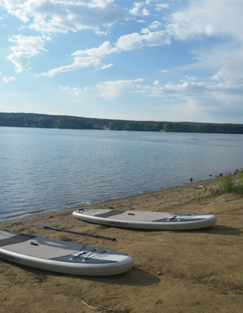 Two paddleboards on a sandy shore by a calm lake under a blue sky with scattered clouds.