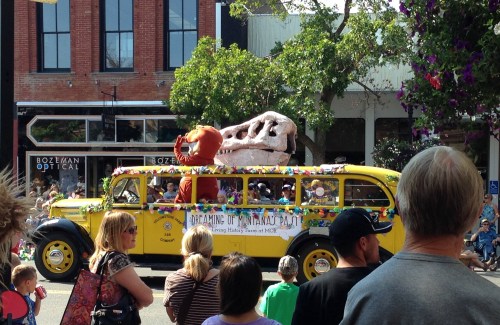 A colorful parade float featuring a dinosaur skeleton and a large dinosaur mascot, surrounded by a festive crowd.