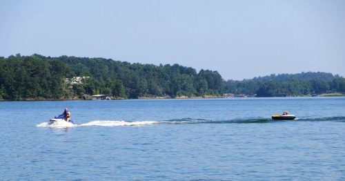 A person on a jet ski tows a small inflatable tube across a calm lake on a sunny day.