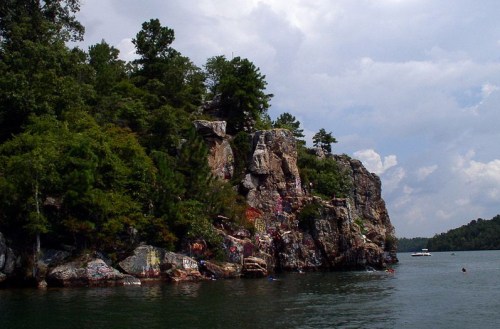 A rocky cliff covered in greenery, with colorful graffiti, rising from a calm lake under a cloudy sky.