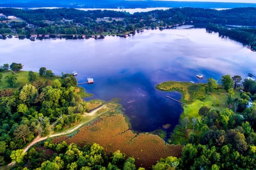Aerial view of a serene lake surrounded by lush greenery and small boats, reflecting the sky and trees.