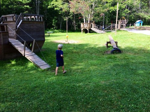 A child walks towards a wooden playground with a pirate ship structure and a bridge in a grassy area.