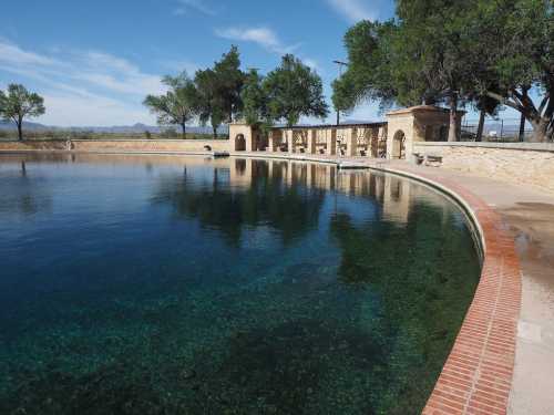 A serene swimming pool surrounded by stone walls and trees, reflecting a clear blue sky.