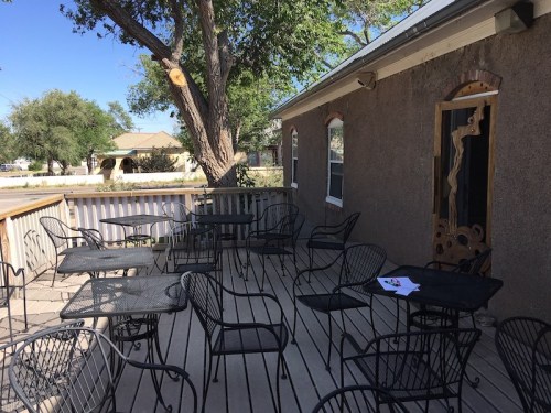 Outdoor patio with empty metal tables and chairs, surrounded by trees and a building under a clear blue sky.