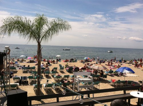 A busy beach scene with umbrellas, lounge chairs, and people enjoying the sun by the water, with boats in the distance.