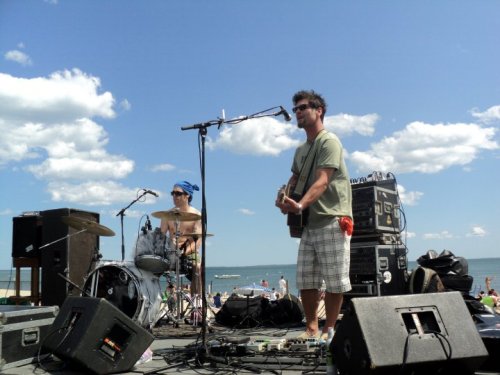 A musician sings and plays guitar on stage at the beach, while a drummer plays beside him under a sunny sky.