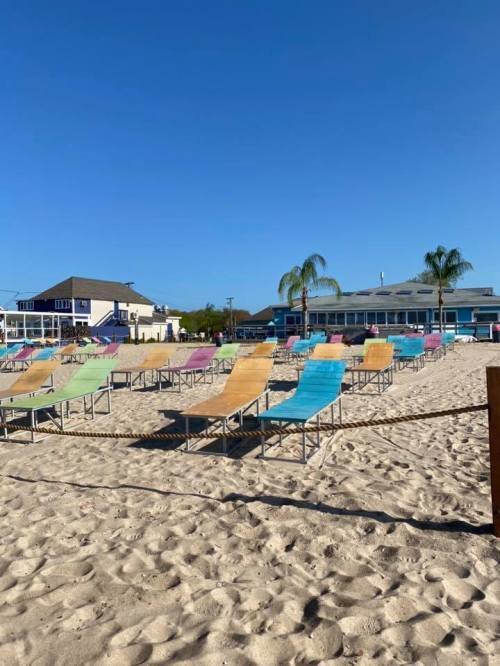 Colorful lounge chairs arranged on a sandy beach under a clear blue sky, with buildings in the background.