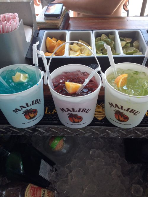 Three colorful cocktails in Malibu-branded buckets, garnished with fruit slices, on a bar counter with ice.