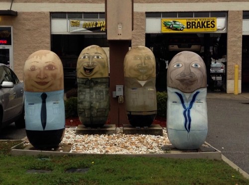 Four large, painted rocks resembling cartoonish people in various outfits, displayed outside a service shop.
