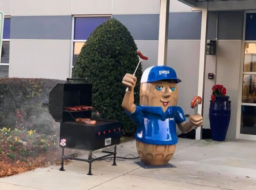 A large peanut mascot in a blue shirt and cap stands by a grill, holding sausages, outside a modern building.