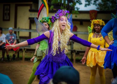 A performer in a purple fairy costume sings joyfully, surrounded by other colorful characters in a lively outdoor setting.