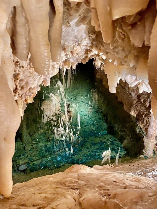 A cave interior featuring stalactites, stalagmites, and a shimmering pool of water reflecting the cave's formations.