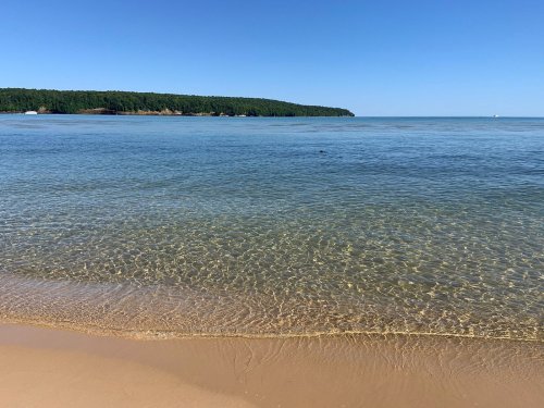 A serene beach scene with clear water, gentle waves, and a forested shoreline under a blue sky.
