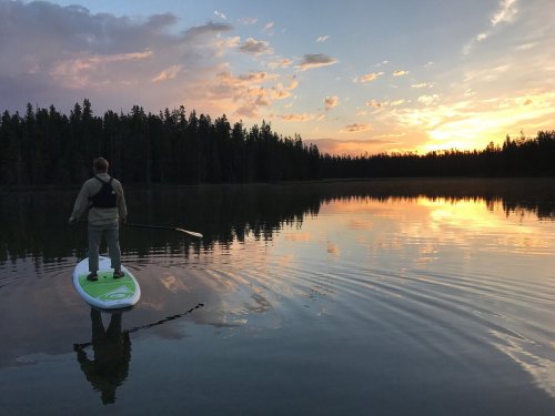 A person paddleboarding on a calm lake at sunset, surrounded by trees and colorful clouds.