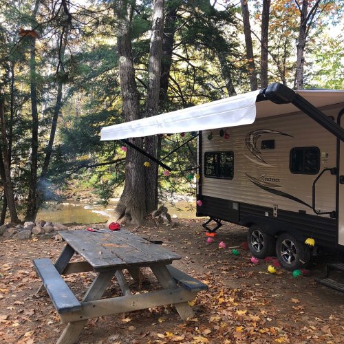 A camper with an awning set up near a picnic table, surrounded by trees and autumn foliage by a river.