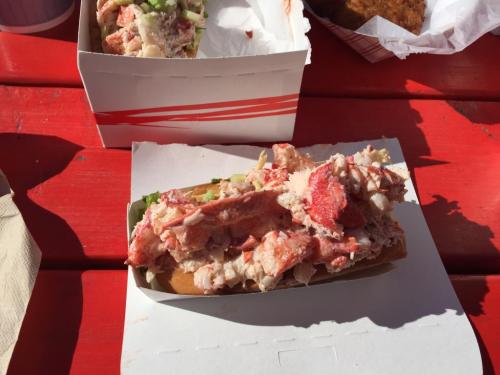 A lobster roll filled with chunks of lobster meat, served on a bun, with a side of fries in a takeout box.