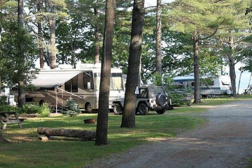 Two RVs parked in a wooded area near a lake, surrounded by trees and a grassy campsite.