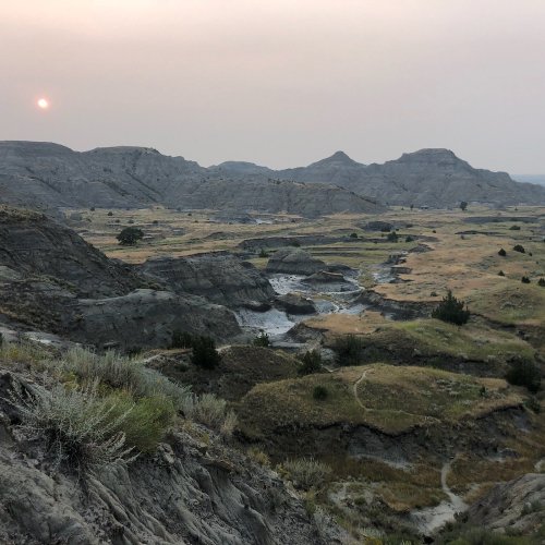 A hazy sunset over rugged hills and a winding river in a dry, grassy landscape.