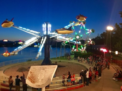 A vibrant amusement park at dusk, featuring a spinning ride and a crowd waiting in front of a lake.