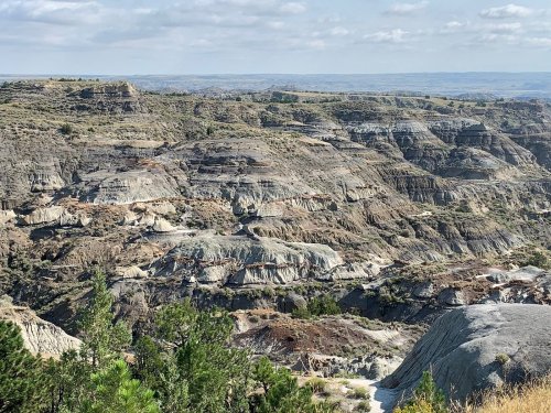 A panoramic view of rugged, layered hills and valleys under a cloudy sky, showcasing natural geological formations.