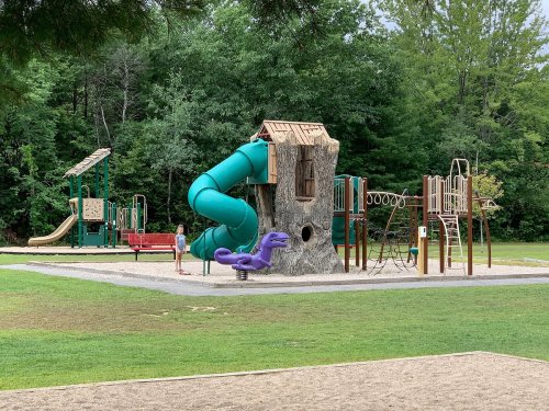 A playground featuring a treehouse slide, climbing structures, and a purple dinosaur sculpture, surrounded by greenery.