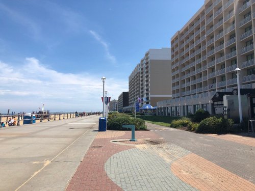 A sunny boardwalk lined with buildings, featuring a path and greenery alongside the beach.