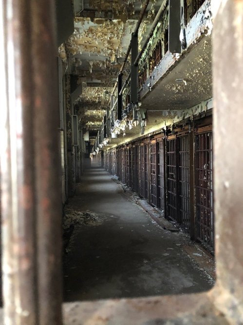 View through a barred window into a dimly lit, abandoned prison corridor with peeling walls and empty cells.
