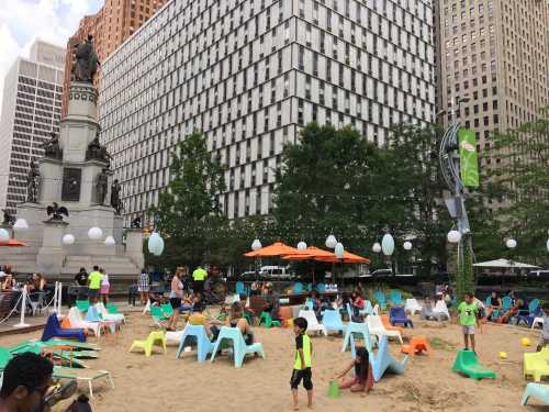 A sandy area in an urban setting with colorful chairs, people relaxing, and children playing with buckets.