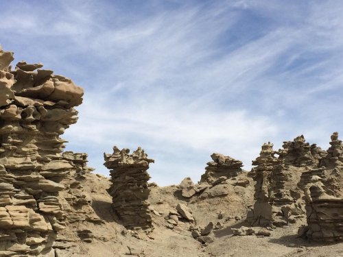 Rock formations rise from a sandy landscape under a blue sky with wispy clouds.