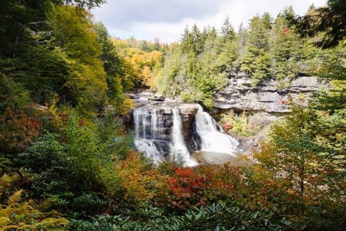 A scenic waterfall surrounded by vibrant autumn foliage and lush greenery under a partly cloudy sky.