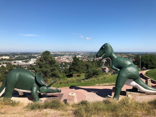 Two large green dinosaur sculptures on a hillside, overlooking a cityscape under a clear blue sky.