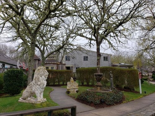 A garden scene featuring stone lion statues, a fountain, and trees, with a house in the background on a cloudy day.