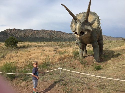 A child stands near a large dinosaur statue in a grassy landscape with mountains in the background.