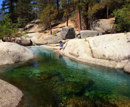 A serene natural pool surrounded by large rocks and trees, with a person wading in the clear water.