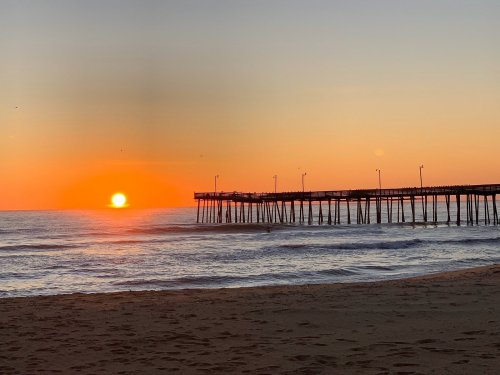 A serene beach scene at sunset, with a pier silhouetted against vibrant orange and yellow skies over calm waves.