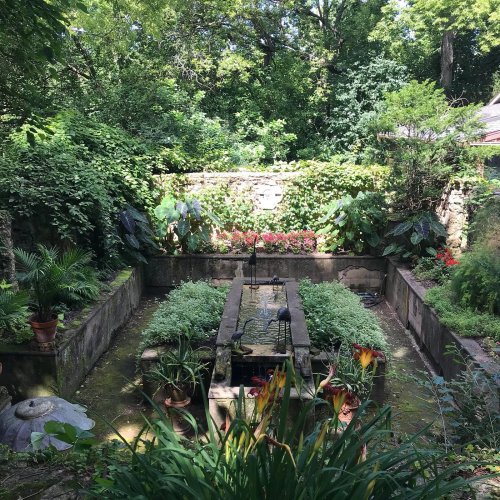 Lush garden with a stone fountain surrounded by greenery and colorful plants in a serene outdoor setting.