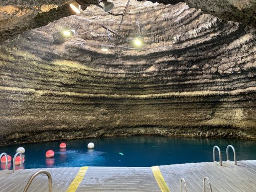 A cavernous space with a deep blue pool, surrounded by rocky walls and floating buoys, illuminated by overhead lights.