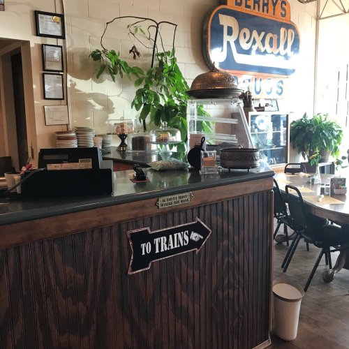 A cozy café counter with a vintage sign pointing to trains, surrounded by plants and a display of baked goods.