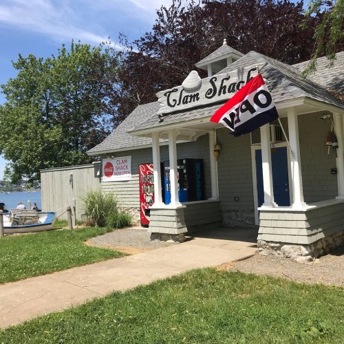 A quaint clam shack with a sign, surrounded by greenery and near a body of water on a sunny day.